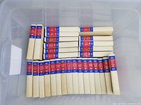 Top-down image showing 29 cream-colored hard cover books stacked neatly inside a plastic container, with red and blue spine labels showing the author's name and book titles.