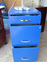 Front view of a blue metal 3-drawer filing cabinet with all drawers partially open, showing interior empty metal drawers and silver handles.