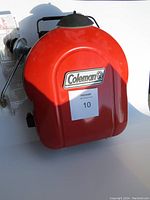 Top view of closed Coleman BBQ grill showing red lid with Coleman logo and a MaxSold lot sticker.