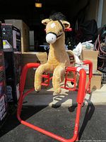Full side view of the plush tan and white hobby horse on red frame with springs and pedals, situated among boxes in a garage or storage area.