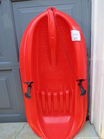 Top view of red plastic snow sled with black steering and brake cables attached on each side, showing molded seat and foot grooves