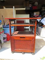 Front view of the wooden printer desk showing the bottom storage compartment with sliding door, open middle shelf, and top shelf surface. Four caster wheels are visible on the base.
