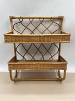 Front view of the two-tier wicker shelf showing the woven lattice back and raised wicker edges around wooden shelf bases.