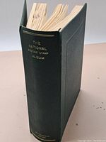 Photo of the large green hardcover album titled 'The National Postage Stamp Album'. The album is thick with visible wear on spine edges and pages.