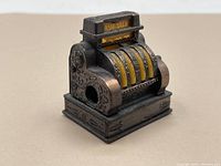 Front-left angle view of the mini pencil sharpener shaped like an antique cash register, showing the insertion hole and decorative engravings