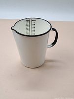 Front and angled view of the white enameled steel measuring cup with black handle and spout, placed on a light surface.