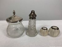 Photo showing two glass containers; a crystal mustard bowl with a sterling silver lid and a crystal sugar shaker with a silver lid, plus two separate sterling silver lids shown separately.