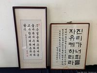 Two framed Chinese calligraphy artworks side by side on a plain wall. The frame on the left (smaller) features vertical black ink characters with a red seal stamp. The frame on the right (larger) has more vertical lines of calligraphy in black ink. Both frames have simple dark wood borders and glass cover. Minor wear visible on frames.