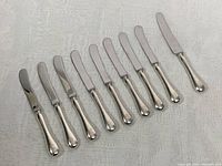 Full view of nine spreader knives arranged in a line, showing silver blades and rounded silver handles.