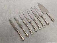 Full view of 7 dinner knives and one cake cutter showing blades and sterling silver handles.