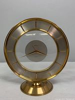 Front view of the entire brass clock with pedestal base, showing clear glass face and gold hour markers.