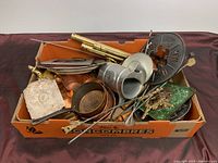 Box of assorted vintage metalware including copper pans, metal plaques with skull designs, candleholders, skewers, and small decorative plates.