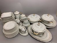 Full view of white porcelain dish set with gold rims including multiple plates, platters, tureens with lids, serving bowls, creamer, and sugar bowl, arranged on gray surface