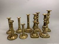 Full group shot showing all 10 brass candleholders of varying designs and heights, including candleholders with round and square bases and different styles of stems.