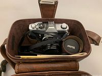 Photo showing open brown leather carrying case containing Canon TLb camera with attached 50mm lens and Vivitar teleconverter lens cap.