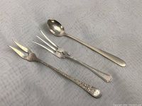 Three vintage sterling silver utensils photographed on white textured fabric: a small spoon, a small fork, and a Birks cocktail fork with ornate handle.