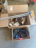Box containing an extendable pole with blue handles, assorted sanding sheets and abrasives, and a separate box of grinding wheels and abrasive discs.