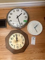 Three wall clocks on wood floor: metal frame clock with black numbers, white plastic clock with raised numbers, wooden octagonal clock with beige face
