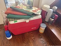 Lot overview showing red Gott cooler, thermoses, water bottle, disposable cups, and folded tablecloths on top and beside cooler on wooden floor.