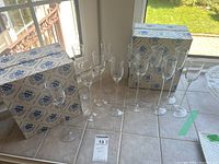 Set of twelve Bereg crystal champagne flutes arranged on a tiled table with two original Bereg branded boxes shown beside them. The flutes are clear and elegant with long stems.
