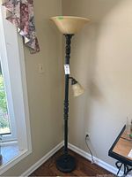 Full view of the floor lamp showing the tall black metal base with carved detailing and the two frosted glass shades, with a window and curtain in the background.