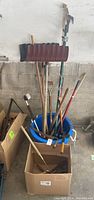 Full view of all garden tools standing in bucket and boxes showing shovels, forks, and pruning shears, and blue plastic bucket.