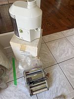Bruan electric juicer in white on a box and Tipo Lusso manual pasta maker with two roller attachments on tiled floor.