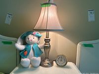 Full view of a table lamp illuminated with beige fabric shade, plush bunny dressed in teal vest and hat, and vintage Home Time analog alarm clock with brass tone framing