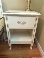 Front view of the vintage nightstand showing drawer and open shelf below, standing on curved legs, white finish with metal drawer pull.