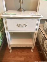 Front view of the white vintage nightstand showing the drawer with decorative metal pull and open shelf below.
