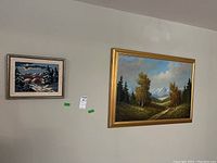 Photo showing both framed items on the wall: a large framed landscape oil painting and a smaller framed needlework.