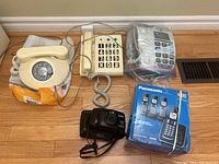 Top-down photo showing the vintage rotary dial phone, two accessible button phones, Panasonic cordless phone in box, and Pentax Espio 738 film camera.