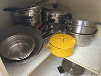 Cookware on two shelves including stainless steel pots, pans, a yellow Dansk saucepan, and stackable bowls