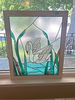 Front view of the stained glass panel showing the swan with surrounding reeds and water in a white wood frame.