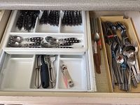 Drawer with plastic compartment trays holding multiple pieces of cutlery including black handled knives, spoons and a collection of forks and spoons in a separate tray.
