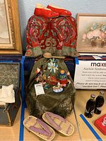 Overview of lot showing red silk jacket draped over object, hand-embroidered shawl with decorative dolls displayed on top, a pair of purple fabric strapped woven wooden geta sandal sandals at front, framed floral painting on right, and a small antique chest on left.