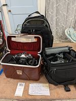 Overall view of lot showing Canon AE-1 Program camera inside vintage brown hard case with red lining, black padded camera bag with extra lenses and straps, and a photographers black backpack in the background.