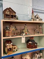Photo of three shelves holding seven ceramic Christmas village buildings including a log cabin house, church, tall clock tower, and shop buildings.