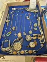 Photo showing full assortment of costume jewelry items laid out on blue fabric inside a cardboard box including necklaces, bracelets, watches and earrings.