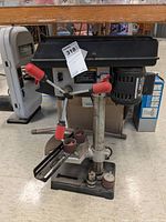 Front view of Craftsman drill press with red handle grips, power cord visible on floor, attached mortising attachment clamps on work table.