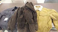 Three jackets laid out on a table: dark brown leather-type jacket, brown leather jacket with shearling collar, and gray quilted jacket.