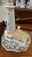 Porcelain lighthouse figurine viewed from angled front showing tower, windows, building with tiled roof and rocky base.