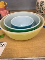 Three Pyrex bowls nested inside each other on a wooden table. Small blue bowl inside medium white bowl inside large yellow bowl. Bowls are vintage with solid colors.