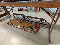 Two Flexible Flyer sleds under a table, one newer with wood and red-metal runners, the other older with rusted metal frame.