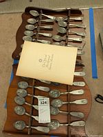 Two wooden racks holding approximately thirteen Franklin Mint official bicentennial commemorative spoons, each decorated with detailed American patriotic scenes and symbols. A certificate/booklet lies on top of the racks.