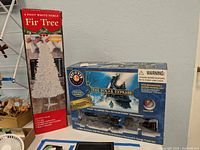 6 ft White Noble Fir artificial tree box and Lionel The Polar Express train set in original box placed side by side against a wall