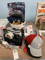 Photo showing a container filled with various sports memorabilia items including a New York Yankees teddy bear, magnet packaging, a leather wallet with cards, and a red Philadelphia Phillies cap alongside a red and gray Phillies cap and a black and white vintage World Series caps.