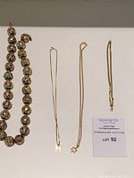 All four necklaces laid out together including multicolor cloisonné bead necklace, two delicate chains with pendants, and box-link chain; auction lot card visible