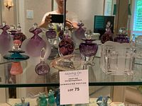 Photo showing multiple glass perfume bottles in lavender, purple, and swirled designs with stoppers, along with present-shaped glass dish, arranged on mirrored shelf.