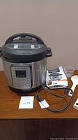 Front and side view of Instant Pot Viva pressure cooker with power cord, user manual, cookbook, two white plastic serving spoons, and clear measuring cup on wooden surface.